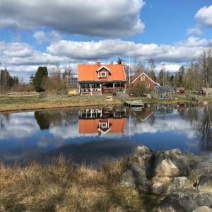 Bild på rött trähus på andra sidan ett vattendrag. Himlen speglar sig i vattnet.