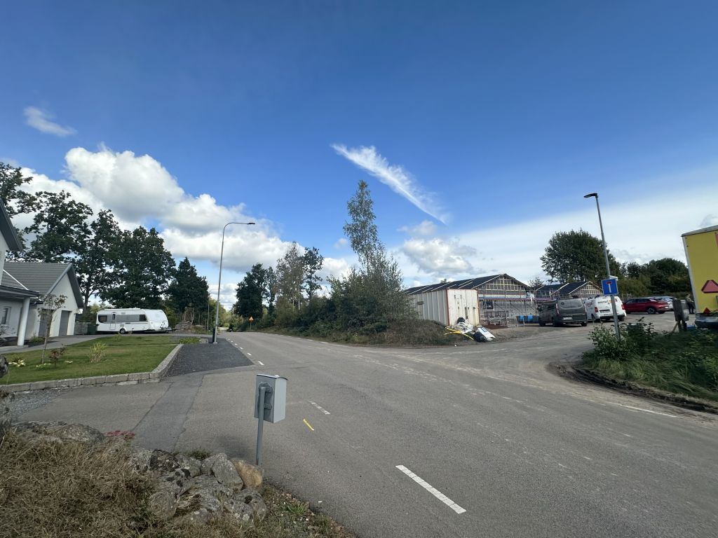 Bild på väg i naturskönt område. Till vänster större vit villa och till höger vägen in till byggplats där hus tar form.